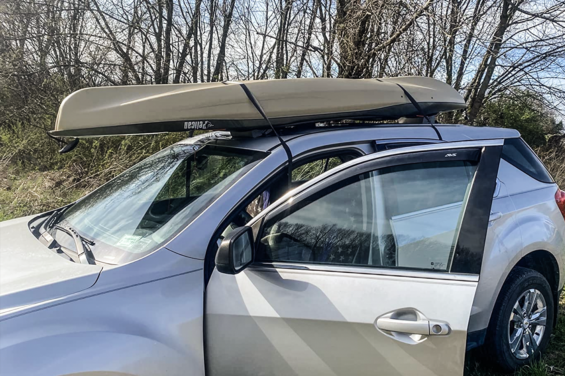A silver SUV with its front door open is parked outdoors, carrying a canoe secured to the roof rack. Leafless trees and grassy vegetation are visible in the background under a blue sky.