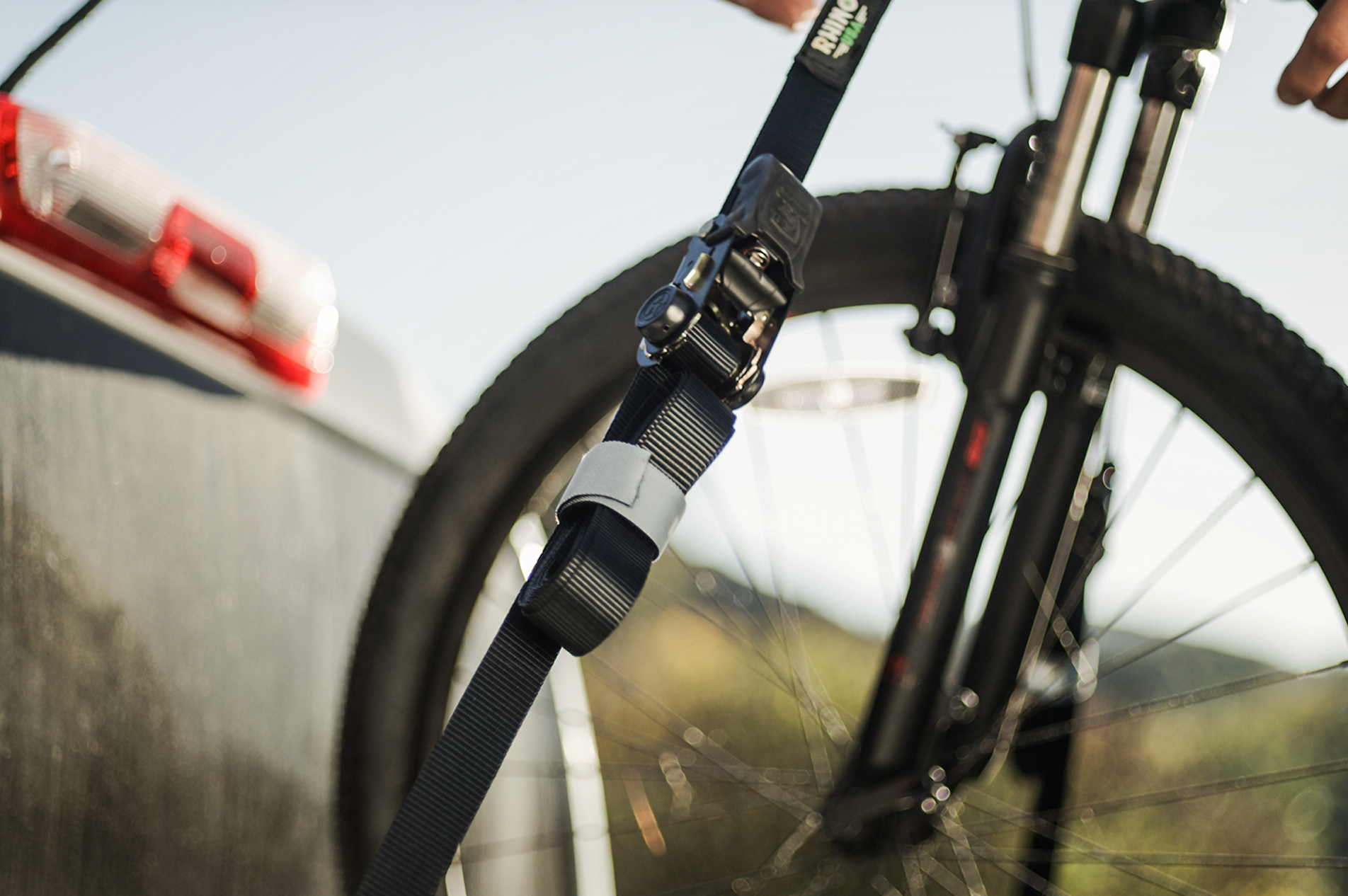A close-up of a bicycle front wheel secured with a black strap and buckle attached to a vehicle, with blurred outdoor scenery in the background.