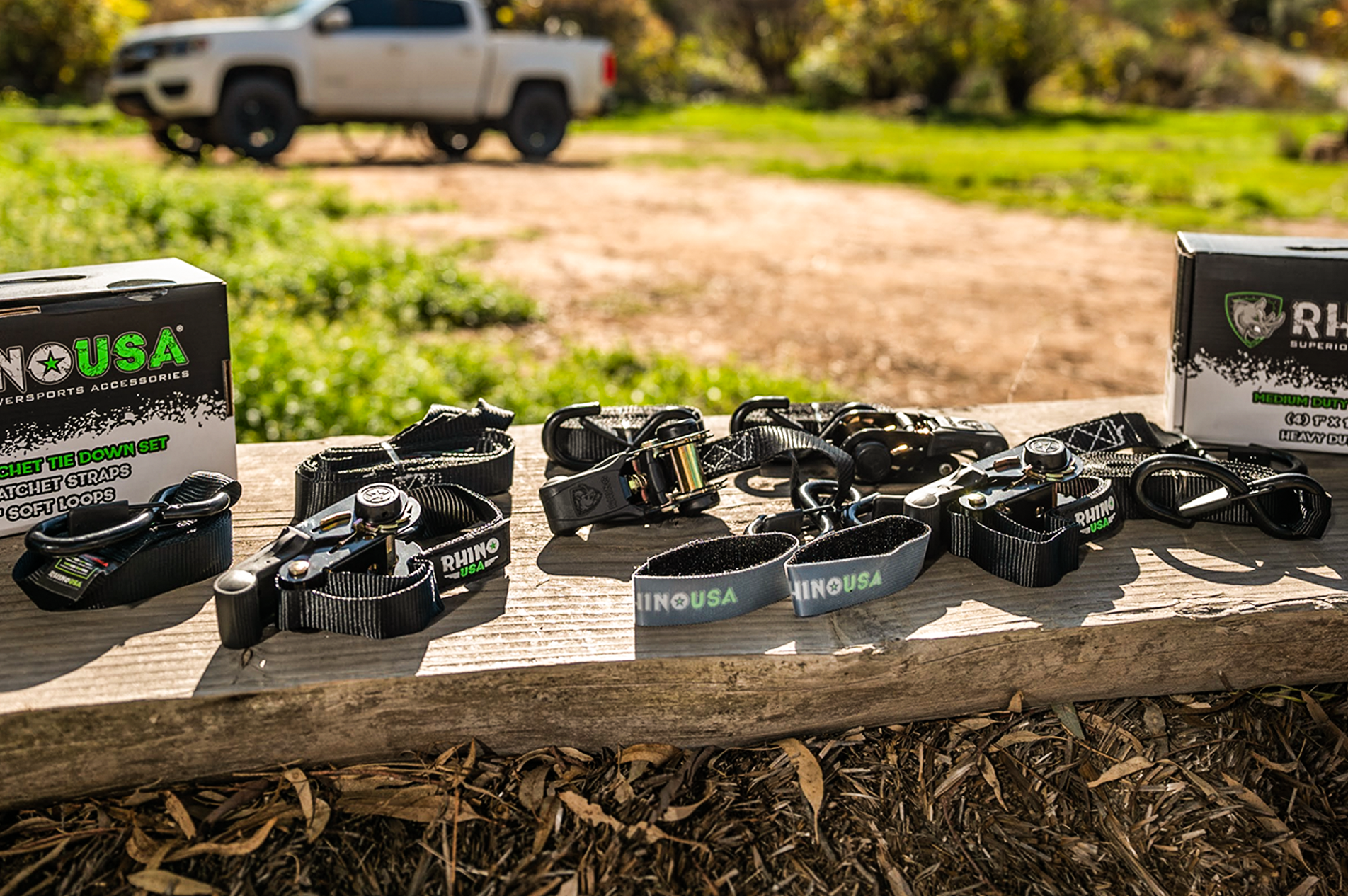 A set of Rhino USA ratchet tie-down straps and accessories are displayed on a wooden plank outdoors, with product boxes on each end and a blurred white pickup truck in the background.